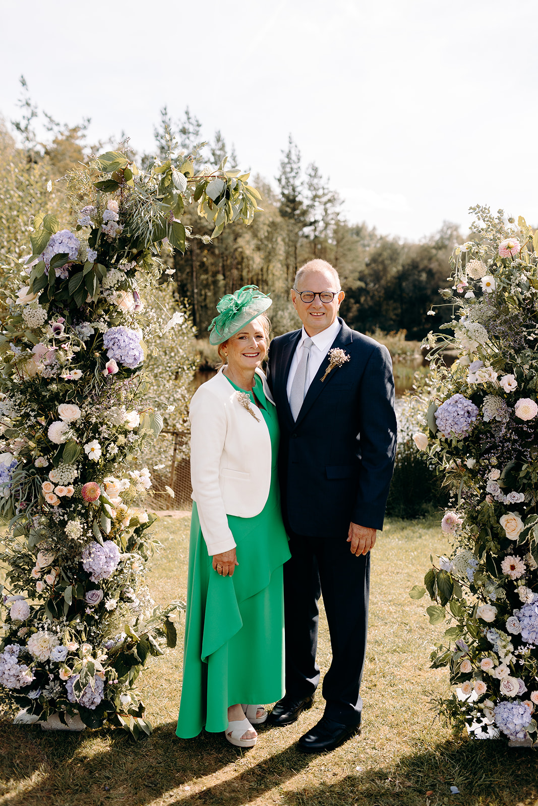 Mother and father of the bride smiling with a stylish guest in front of floral arch, relaxed Darwin Lake group portrait.