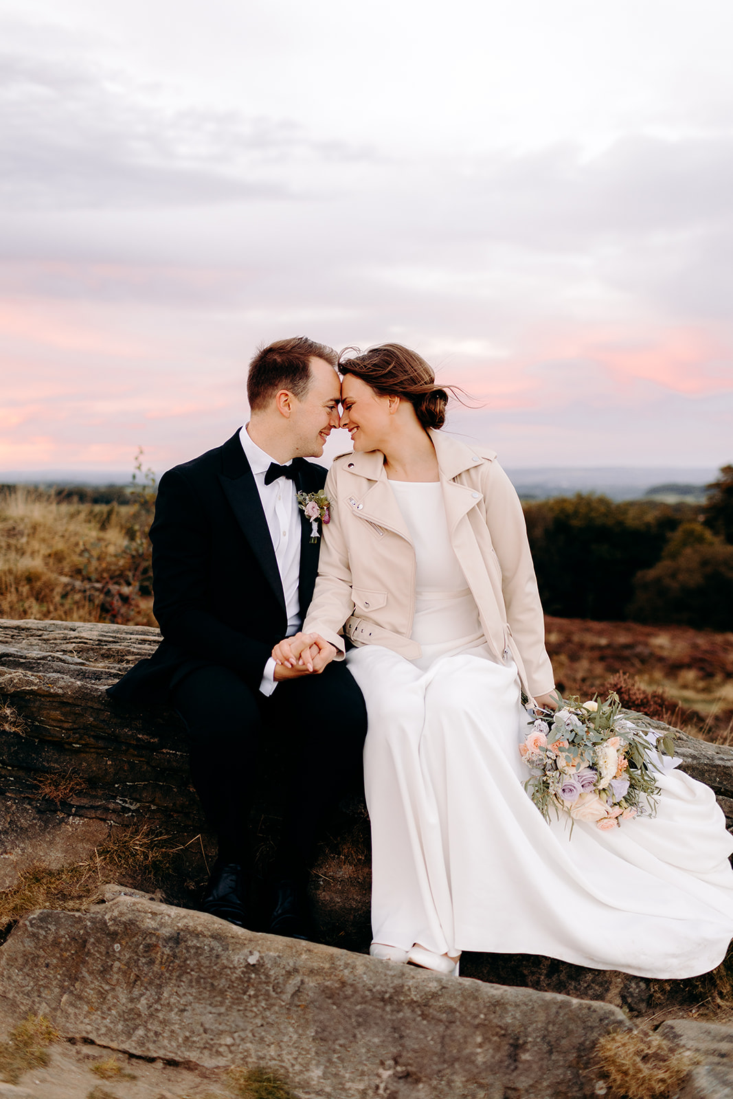 Bride’s dress draped elegantly over moorland stone as she sits with bouquet – editorial style wedding detail.