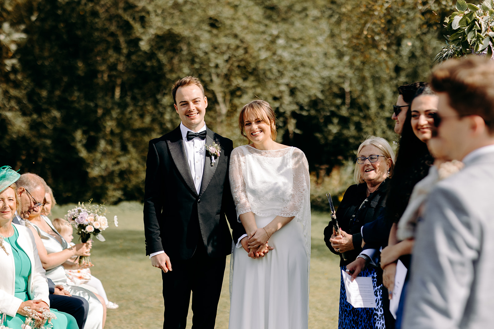 Bride and groom smiling whilst listening to their friends do a reading in their outdoor wedding ceremony.