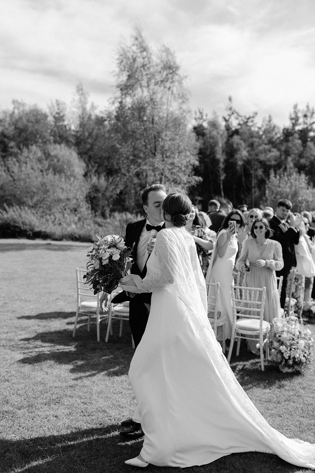 Black and white photo of bride walking away from ceremony area holding train, captured in a candid natural style.