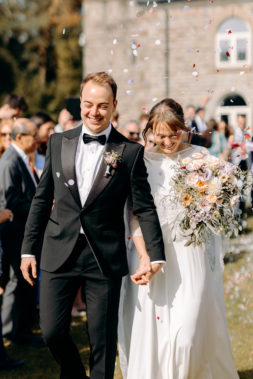 Couple holding hands, mid-confetti tunnel, with colourful paper fluttering around them.