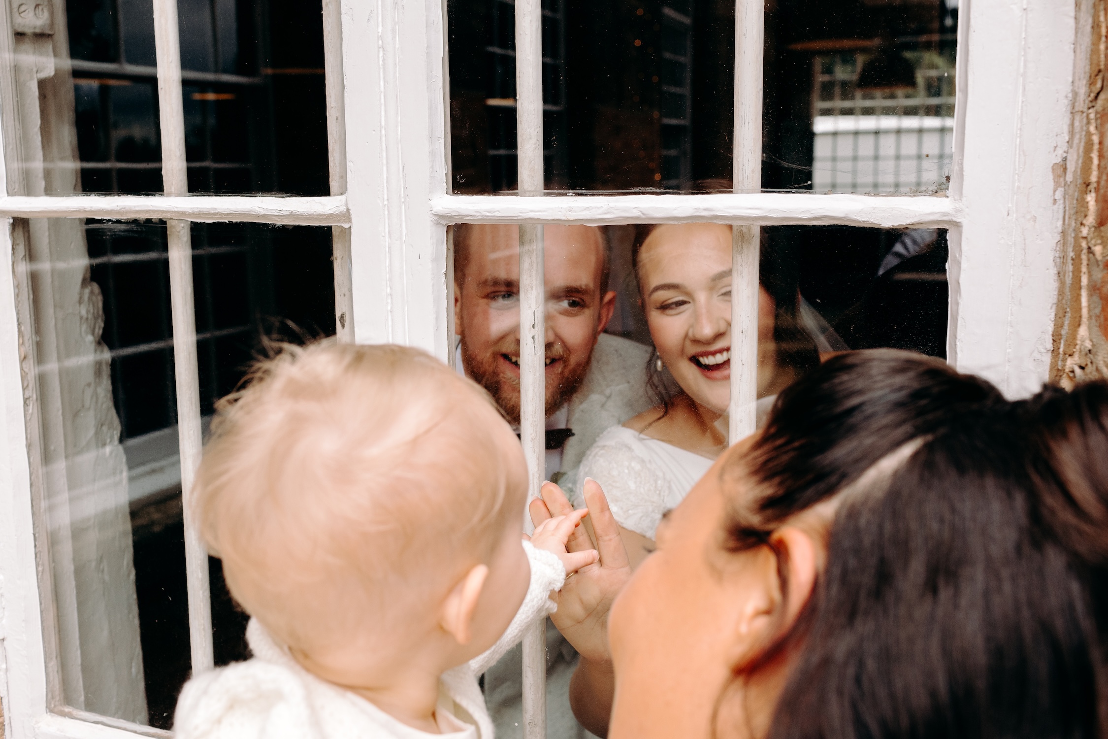 Bride and groom waving to their daughter through the window in this natural wedding photo at The West Mill
