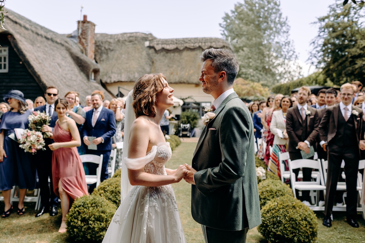 A wide view of the garden ceremony shows guests seated on both sides, smiling as the couple begins their vows – sunny outdoor wedding at Marleybrook House.