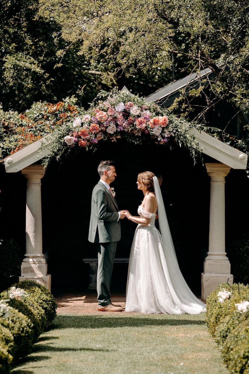 Groom stands confidently beneath the floral arch, reading aloud with a warm expression – personal vows during garden ceremony at Marleybrook House.