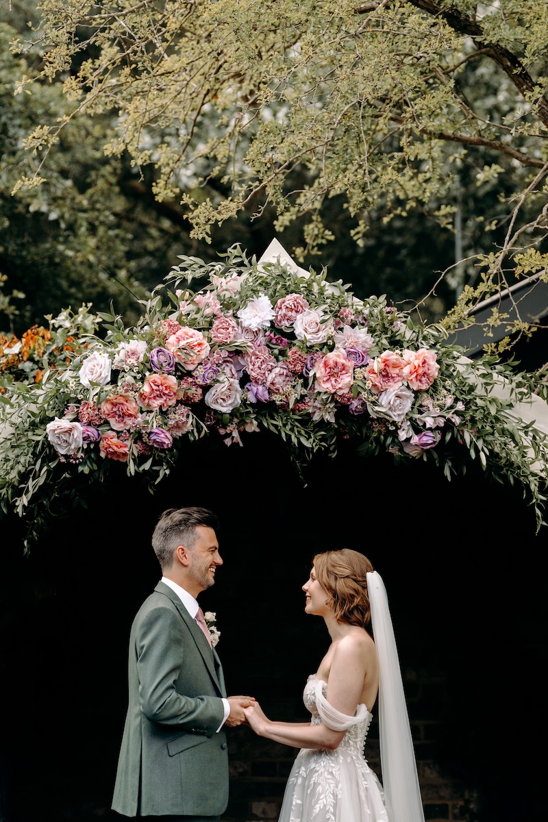Couple holds hands and exchanges a quiet smile as they stand under the archway – natural, emotion-led wedding photography at Marleybrook House.