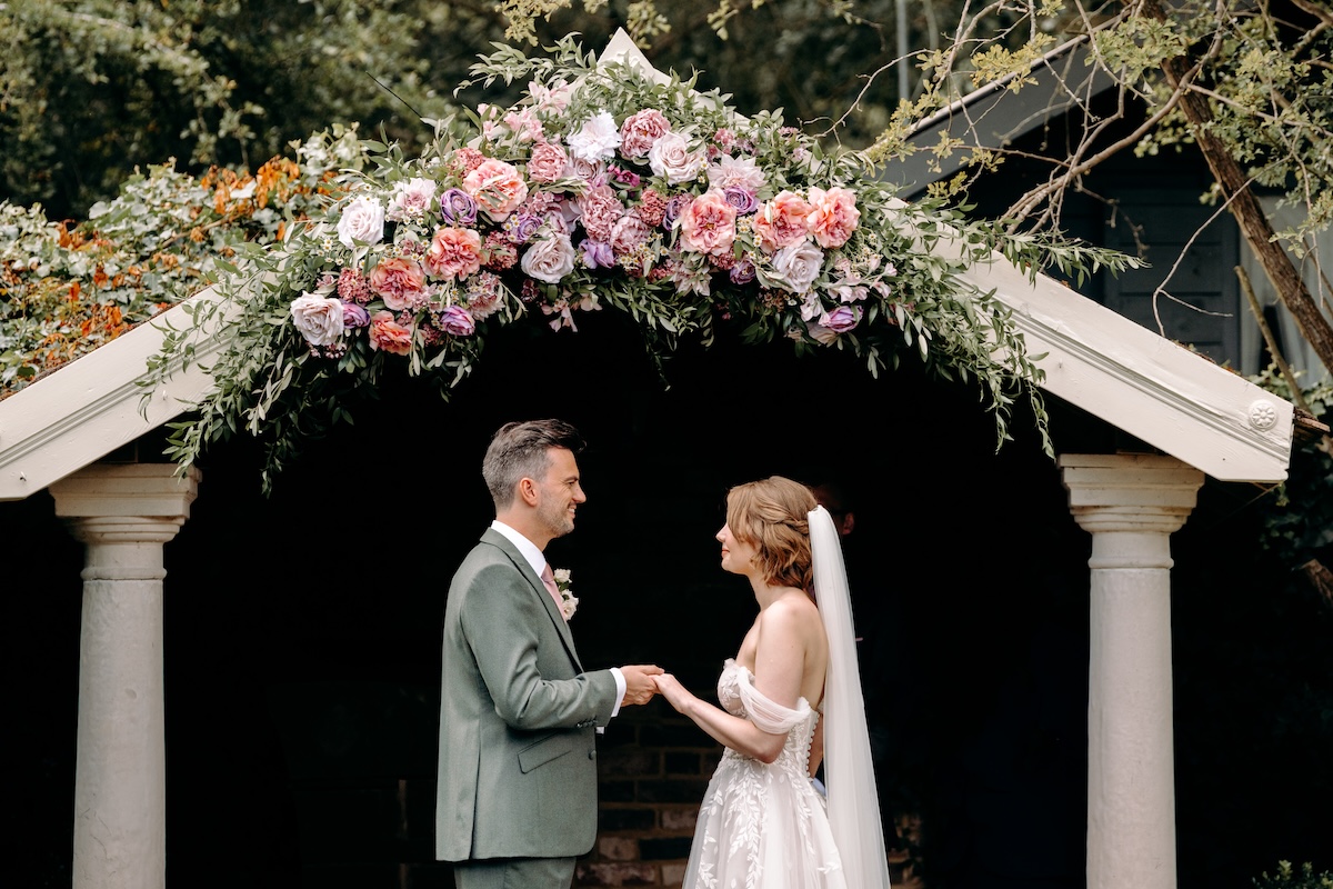 The garden ceremony space at Marleybrook House, with the couple centered under the pink floral arch and guests smiling in the sunshine.