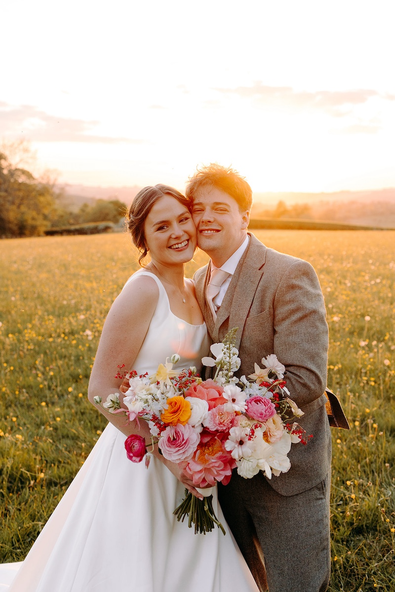 Bride and groom smiling with countryside behind them, bathed in warm evening light.