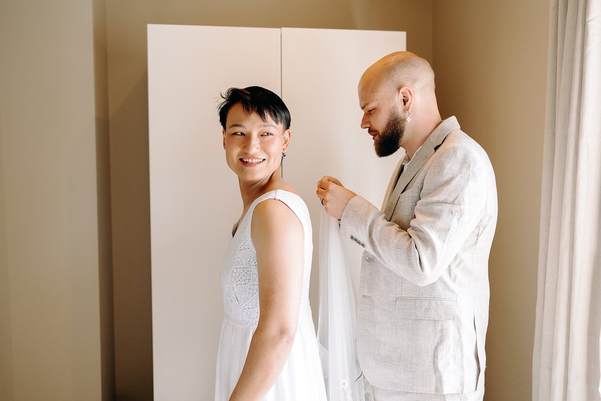 Bride smiling as her partner fastens her dress near a window during a relaxed Peak District elopement morning.