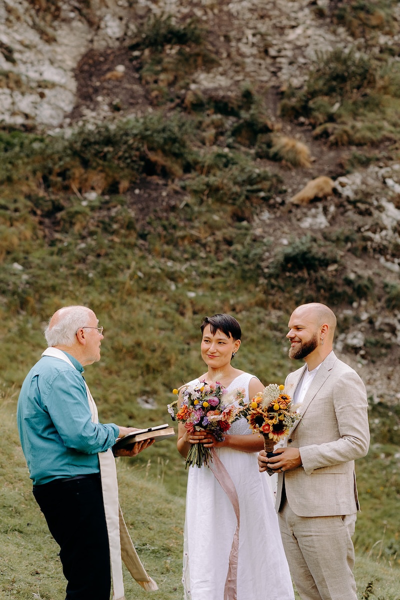 Couple standing together holding flowers beneath tall limestone cliffs at their Peak District elopement.