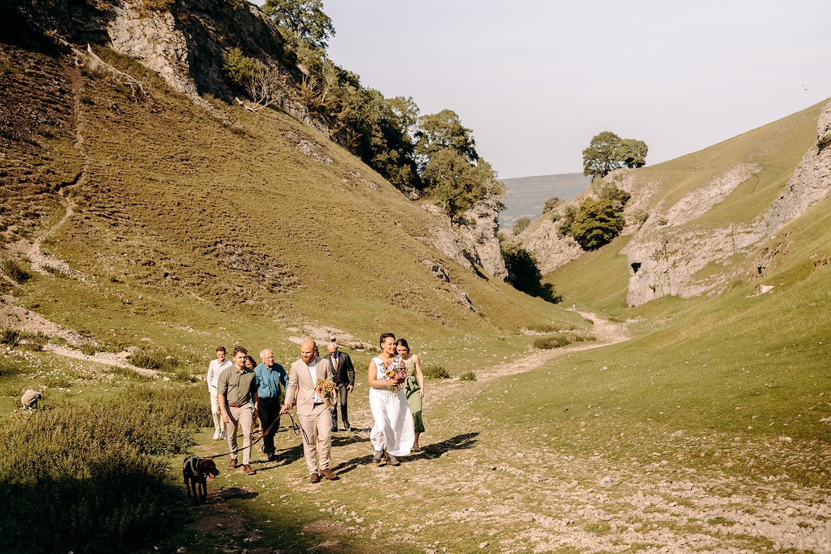 Couple walking through a grassy valley during their Peak District elopement with family and dogs.