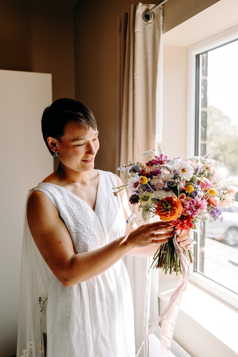 Bride holding a colourful bouquet by the window during their nature-inspired Peak District elopement.
