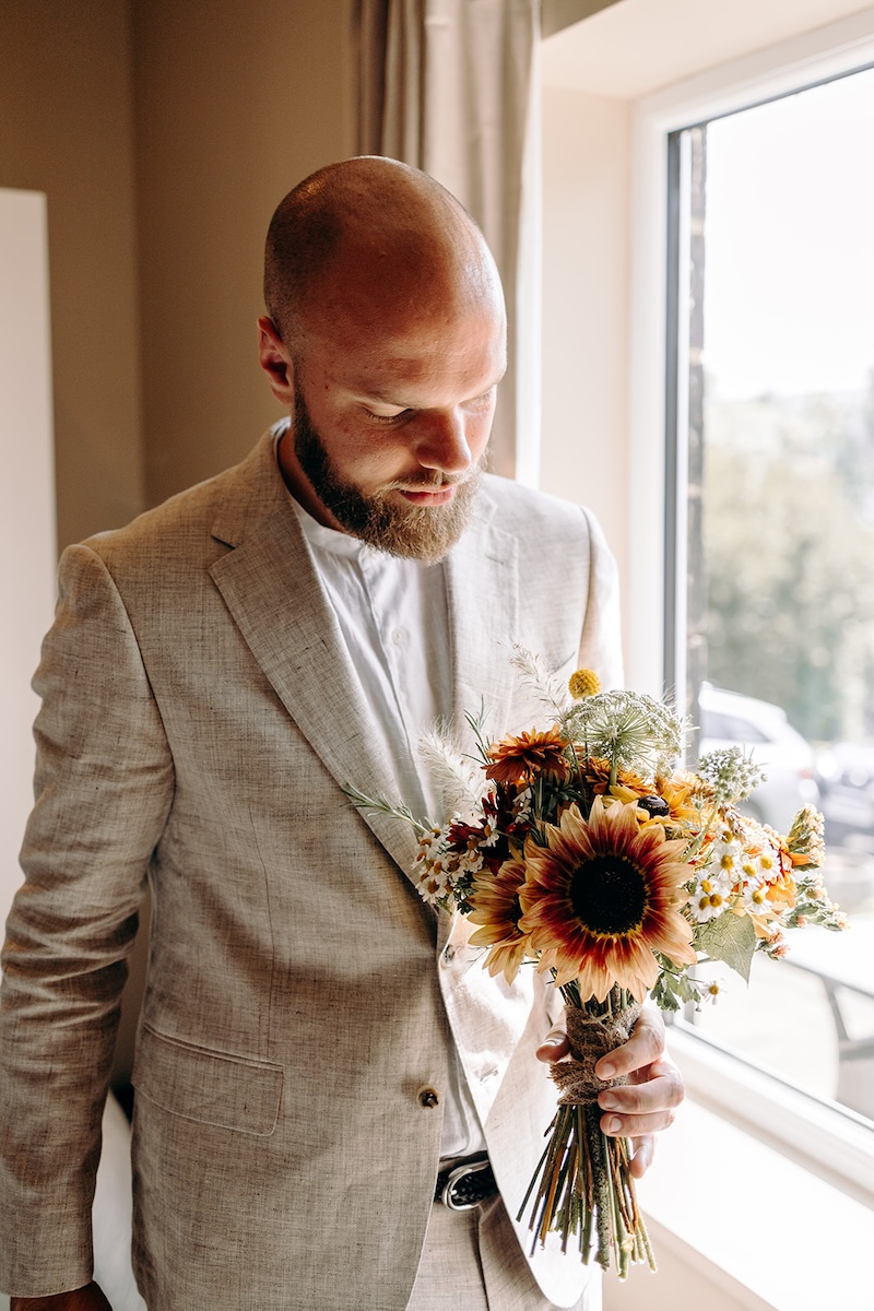 Groom standing by the window holding the sunflower bouquet during their intimate UK elopement.