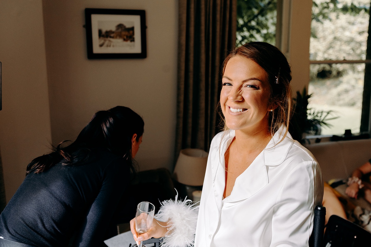 Bride getting makeup done by the window, smiling softly during bridal prep at Eyam Hall – natural Derbyshire wedding photography.
