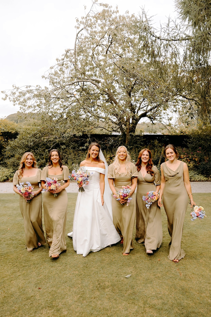 Bride with bridesmaids in pastel dresses smiling together in Eyam Hall gardens – Peak District wedding photographer.