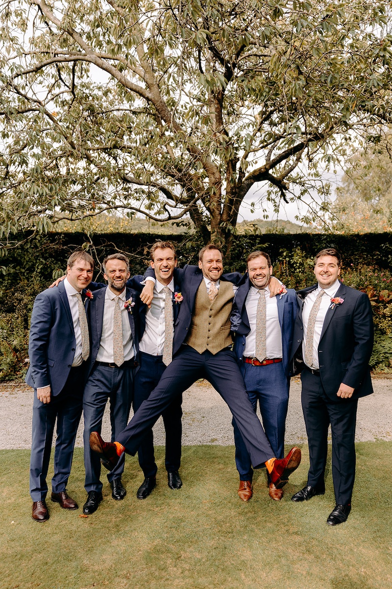 Fun groomsmen group photo under trees in the grounds of Eyam Hall – relaxed natural wedding photography.