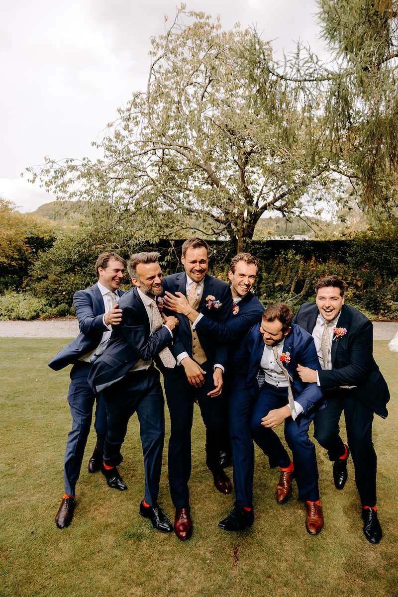 Fun groomsmen group photo under trees in the grounds of Eyam Hall – relaxed natural wedding photography.