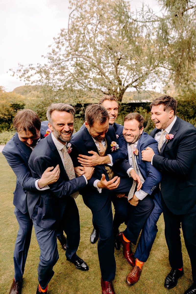 Fun groomsmen group photo under trees in the grounds of Eyam Hall – relaxed natural wedding photography.