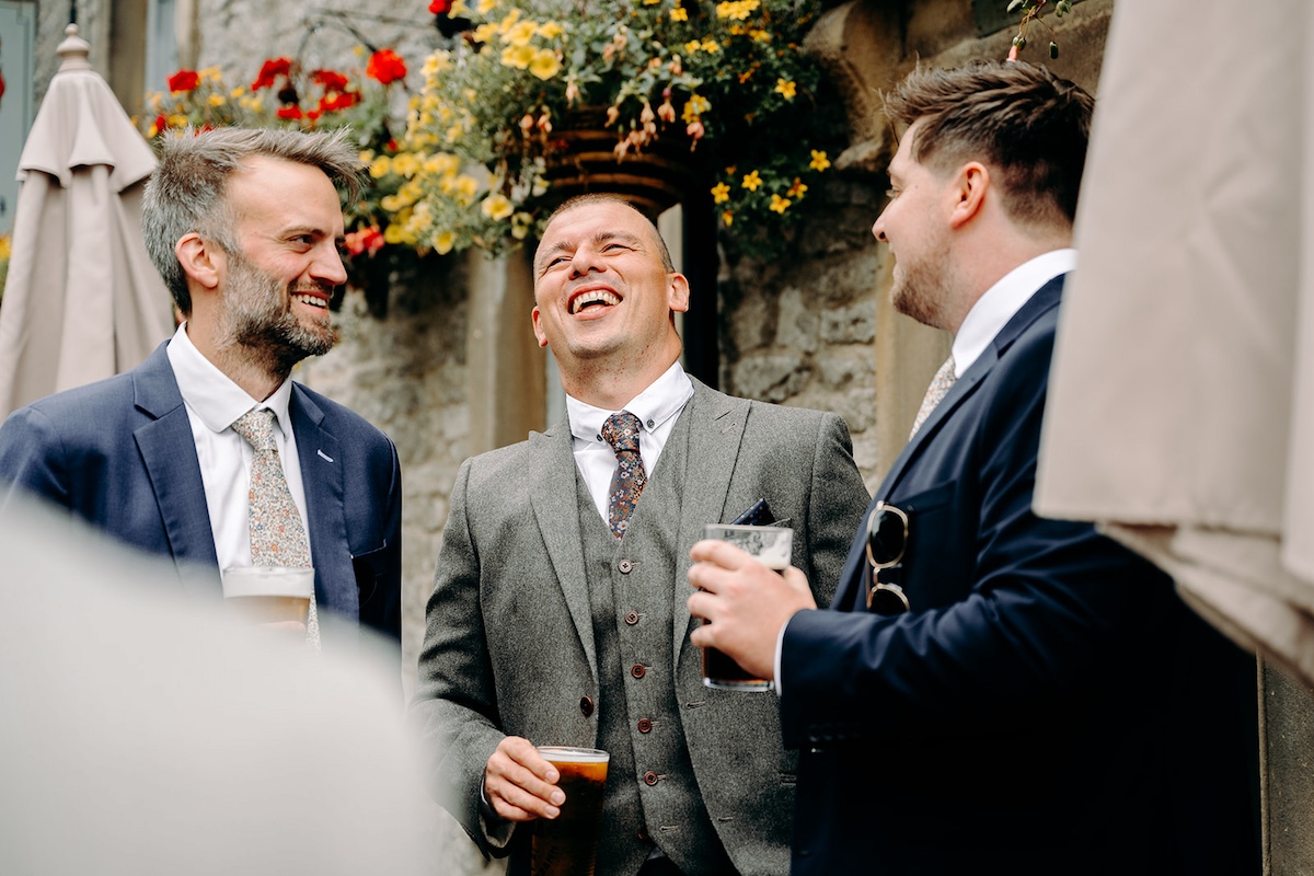 Groom and wedding party stand together laughing outside Eyam Hall church before ceremony.