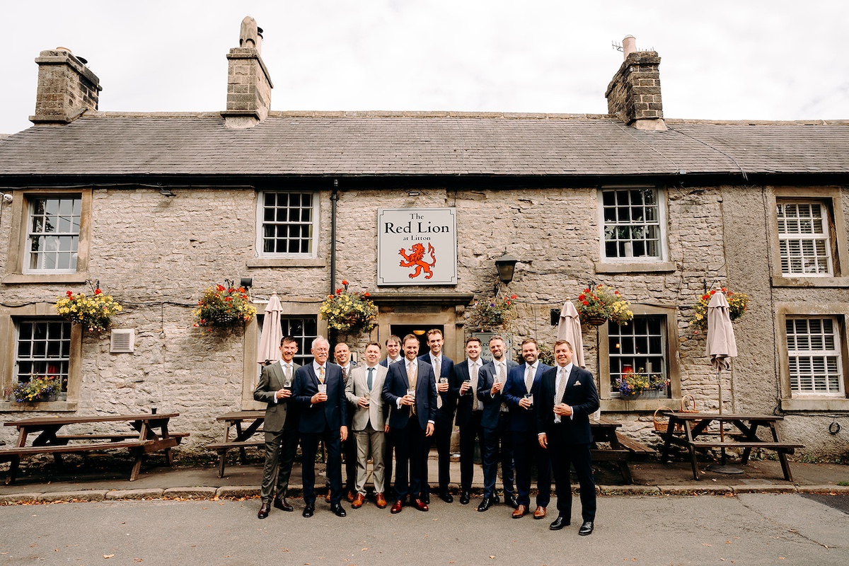 Groom and best man standing together laughing outside Eyam Hall church before ceremony.
