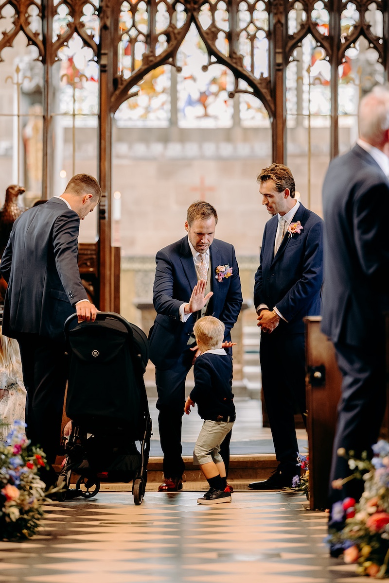 Groom standing at altar high fiving his nephew as ceremony begins inside Eyam Hall church.