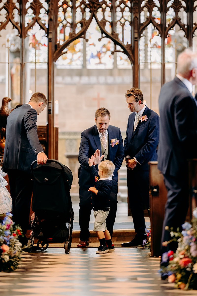 Groom standing at altar high fiving his nephew as ceremony begins inside Eyam Hall church.
