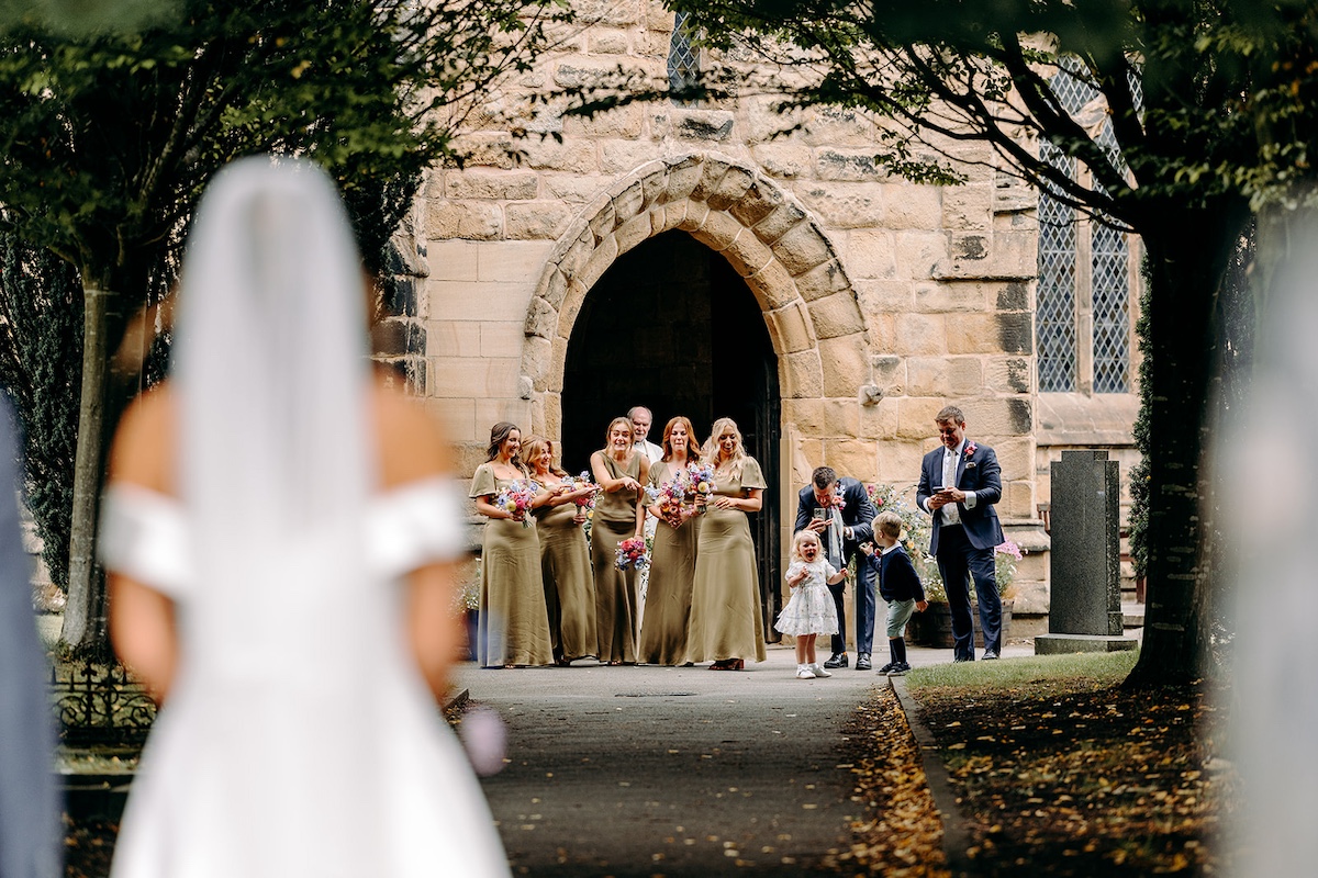 Bride arriving for Peak District church wedding, as bridesmaids and flower girl see her. Natural wedding photography.
