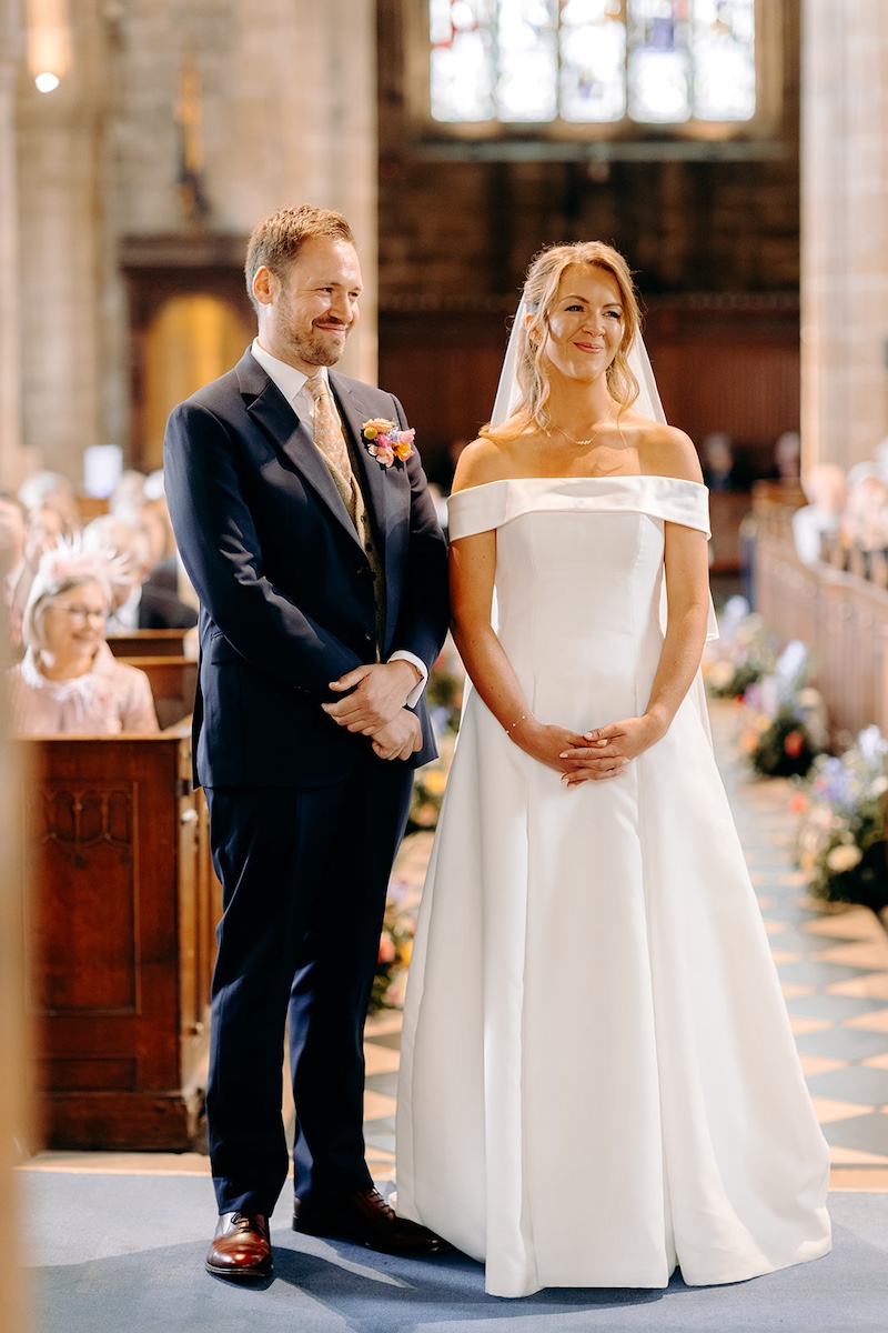 Bride and groom smiling nervously while holding hands during ceremony at Eyam Hall church.