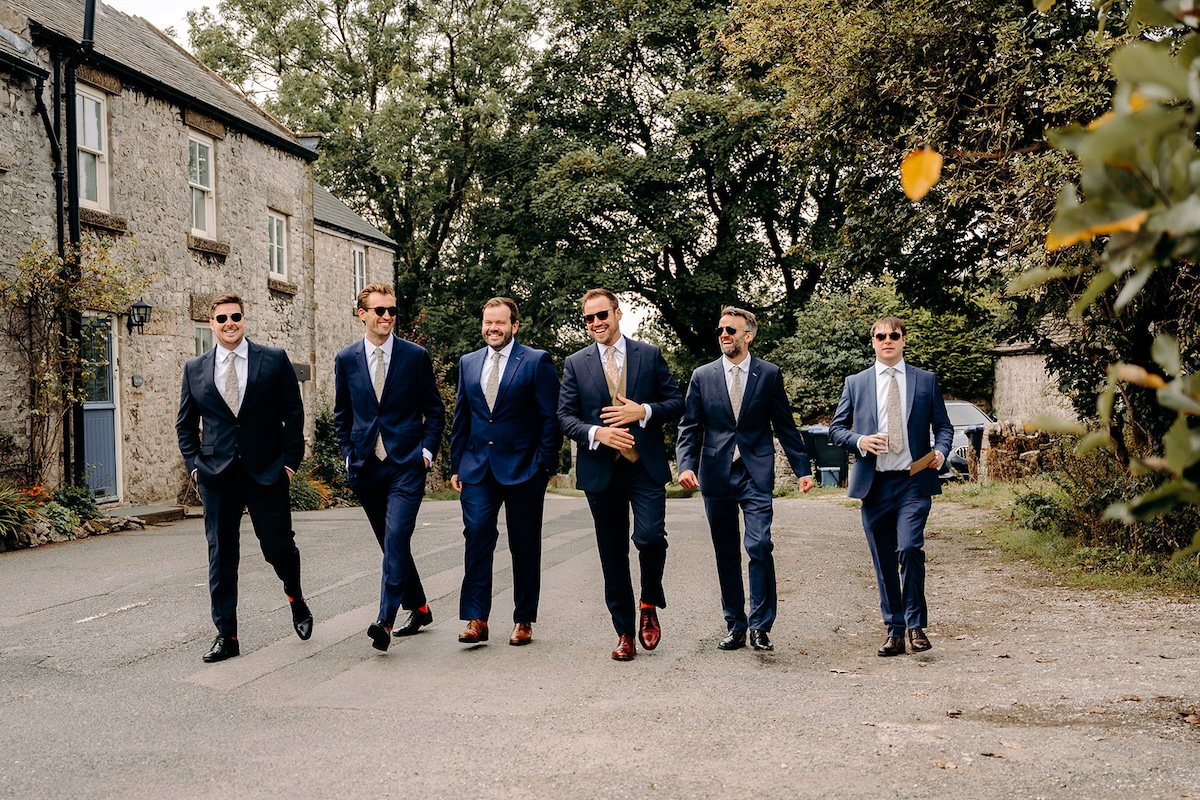 Groom and groomsmen walking through Eyam village in navy suits before the ceremony – Peak District wedding photographer.