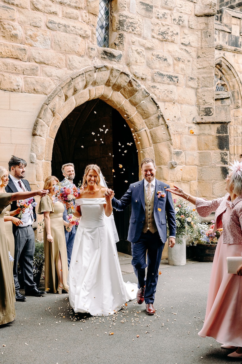 Bride and groom walking out of Eyam church beneath trees while guests throw confetti – joyful Derbyshire wedding photography.