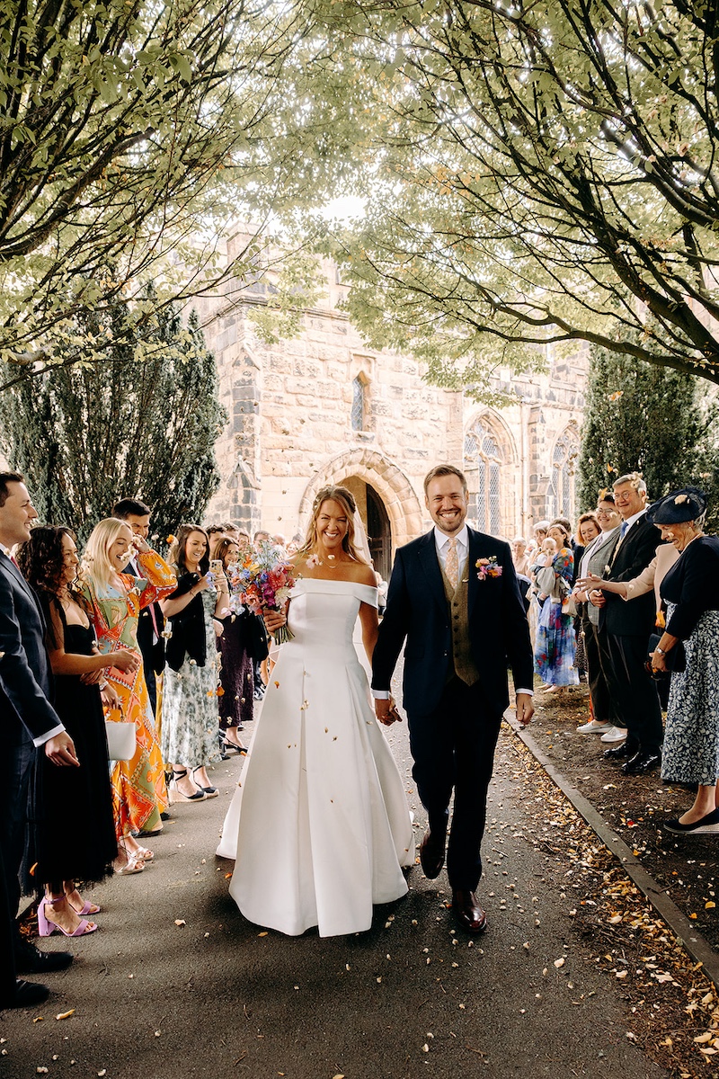 Just-married couple smiling and holding hands under avenue of trees outside Eyam church in the Peak District.
