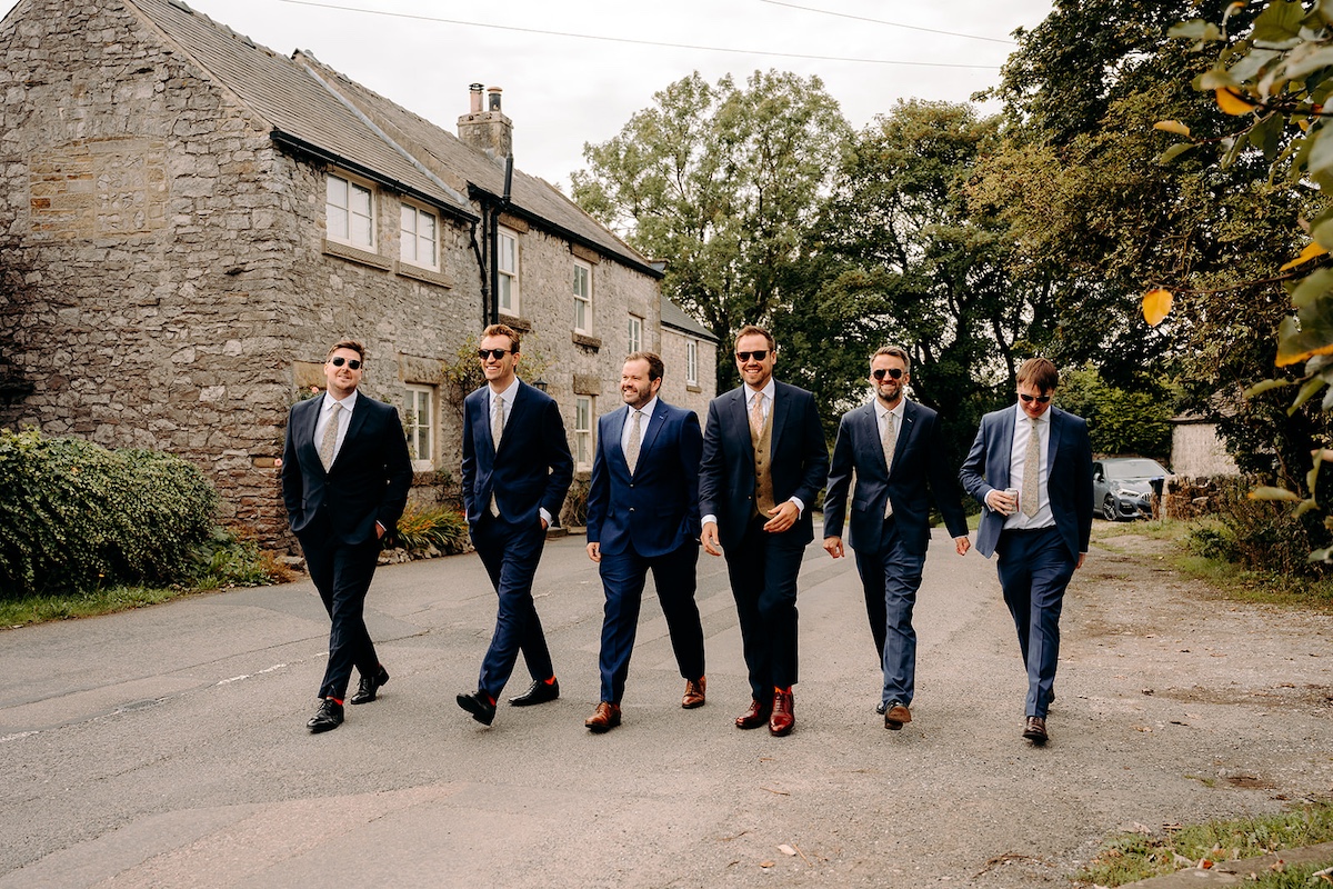Groom and groomsmen walking through Eyam village in navy suits before the ceremony – Peak District wedding photographer.