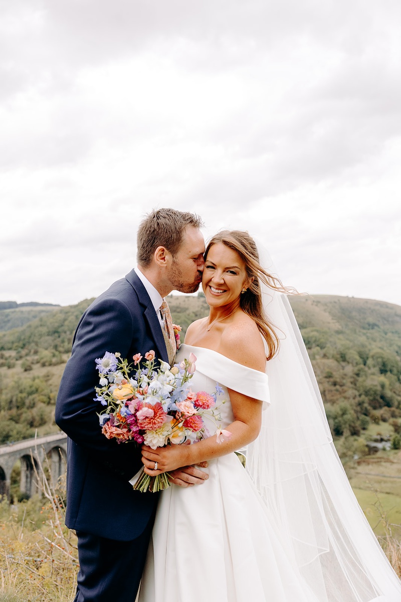 Groom kisses bride’s cheek while she smiles, scenic hills behind – Eyam Hall wedding portraits.