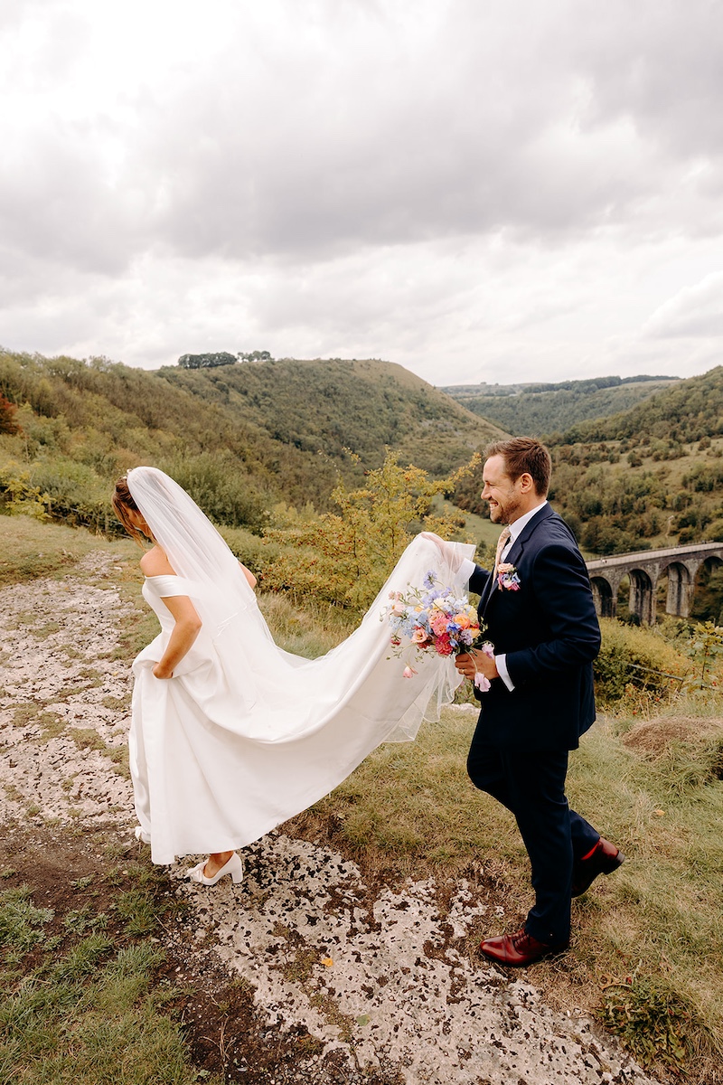 Couple laughing together while bride lifts wedding dress to walk through moorland grass – natural Peak District wedding photographer style.