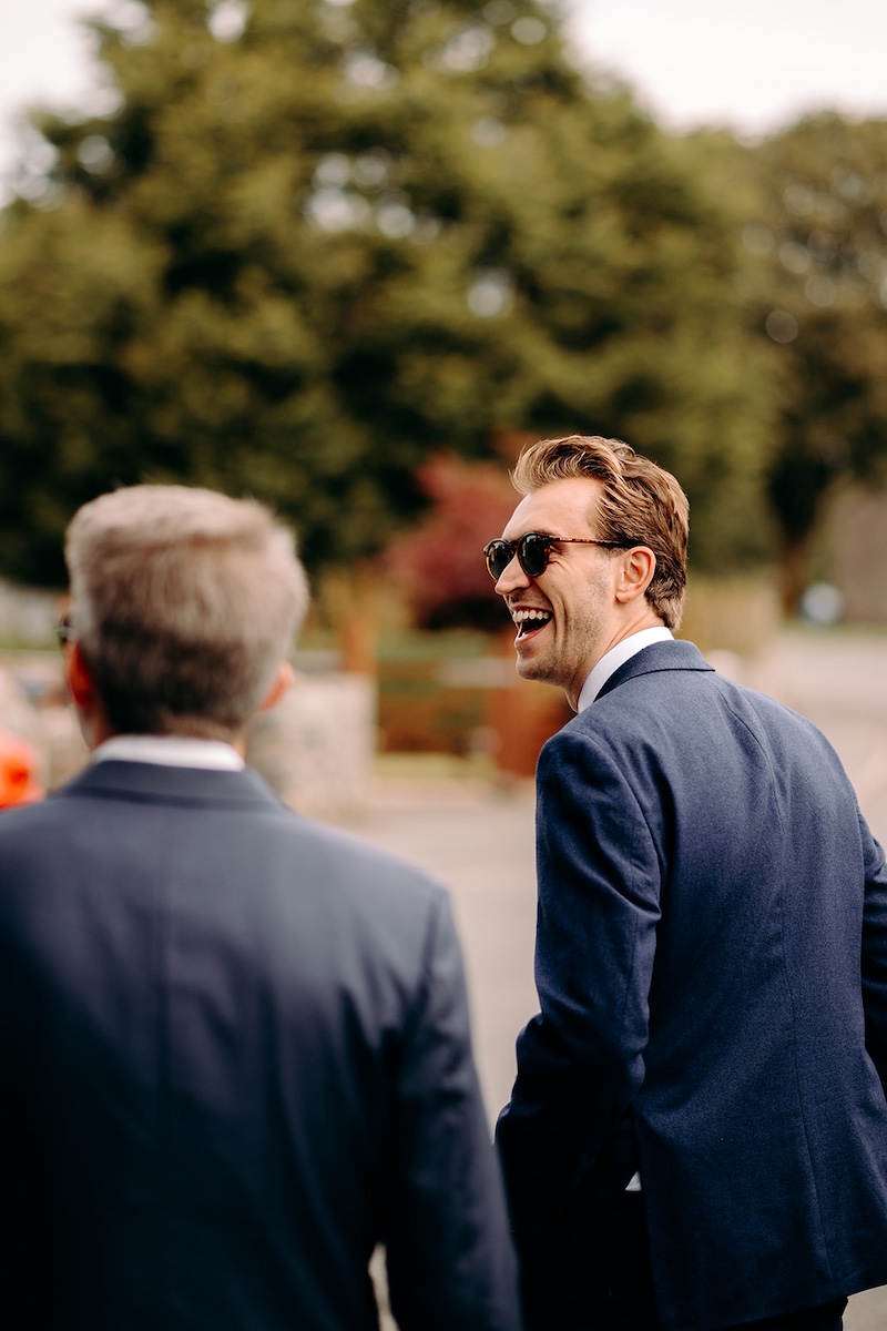 Group photo of groomsmen laughing together outside Eyam Hall church before the wedding ceremony.