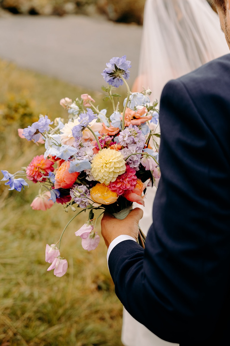 Intimate close-up of groom holding the bride's natural, pastel coloured wedding bouquet as they walk across the Peak District moorlands.