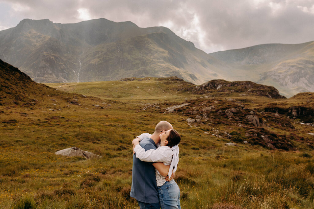Couple cuddling during pre-wedding shoot in North Wales with sweeping mountain views behind them