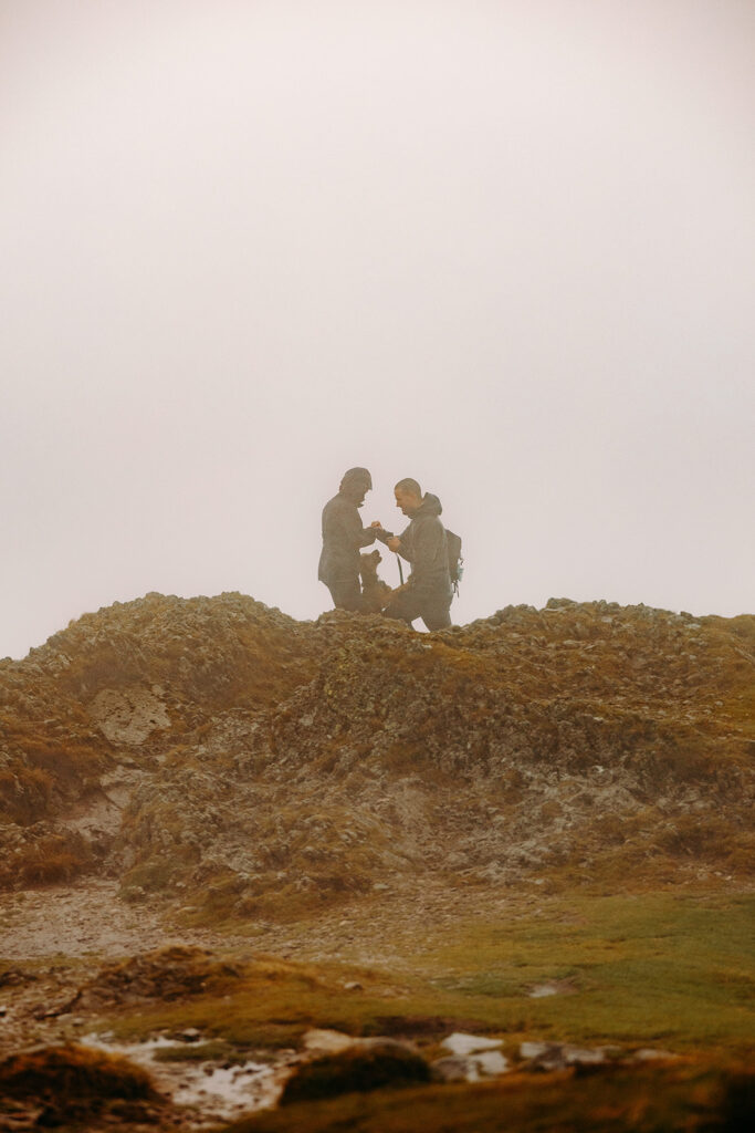 Golden light silhouette of engaged couple and their dog celebrating secret proposal on Lake District mountain ridge