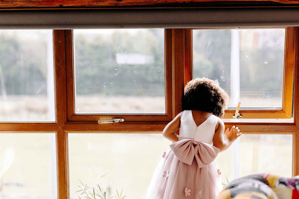 Young flower girl standing by the window in a pink dress during wedding preparations at The Wellbeing Farm, captured with natural documentary wedding photography.