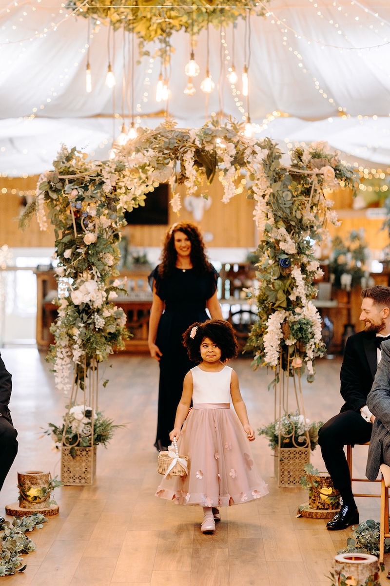 Flower girl walking down the aisle during the ceremony at The Wellbeing Farm, photographed with joyful documentary wedding photography.