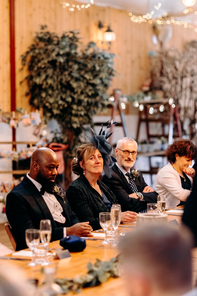 Wedding guests listening to speeches at long reception tables inside The Wellbeing Farm barn.