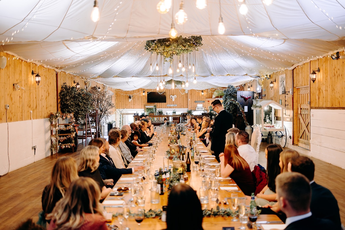 Wide view of the decorated barn reception with guests seated at long tables, photographed by a documentary wedding photographer.