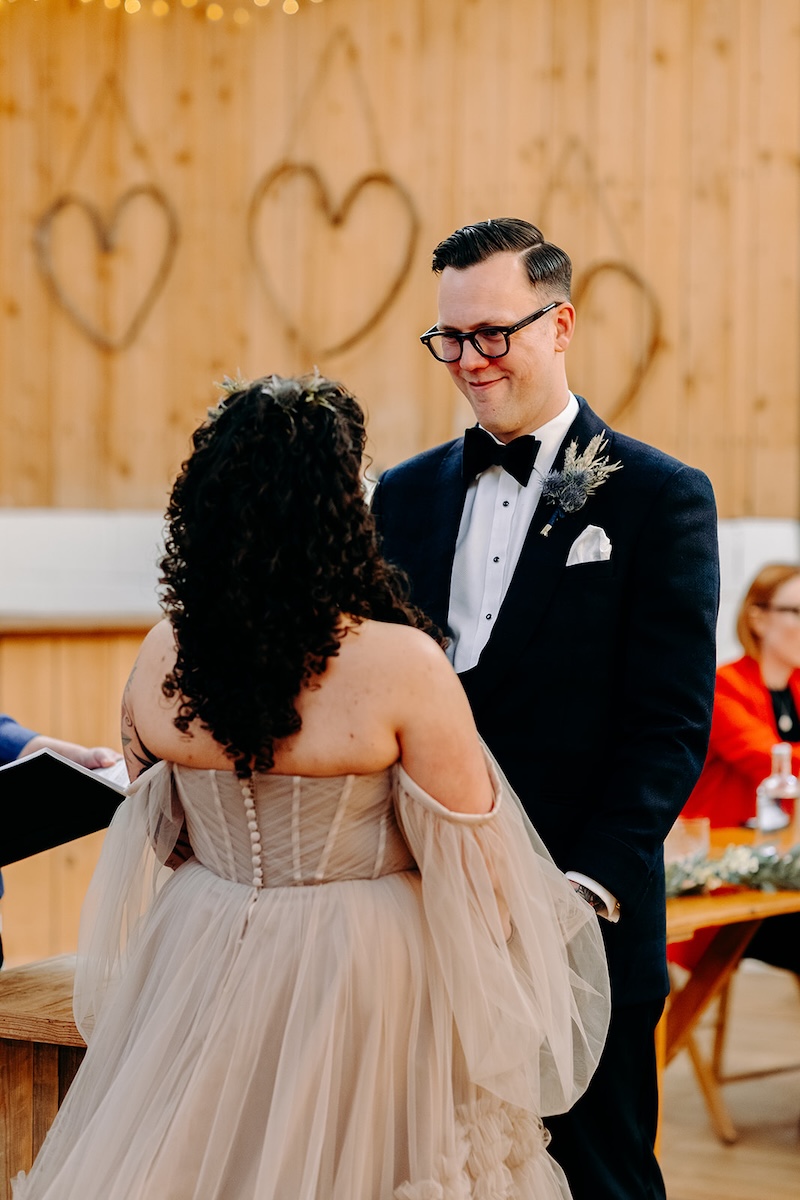 Groom smiling at the bride during their ceremony in the barn at The Wellbeing Farm, captured with relaxed wedding photography.