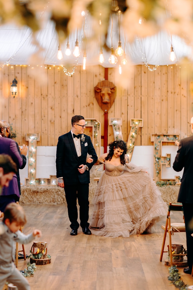 Bride and groom smiling while guests applaud inside the barn ceremony space.