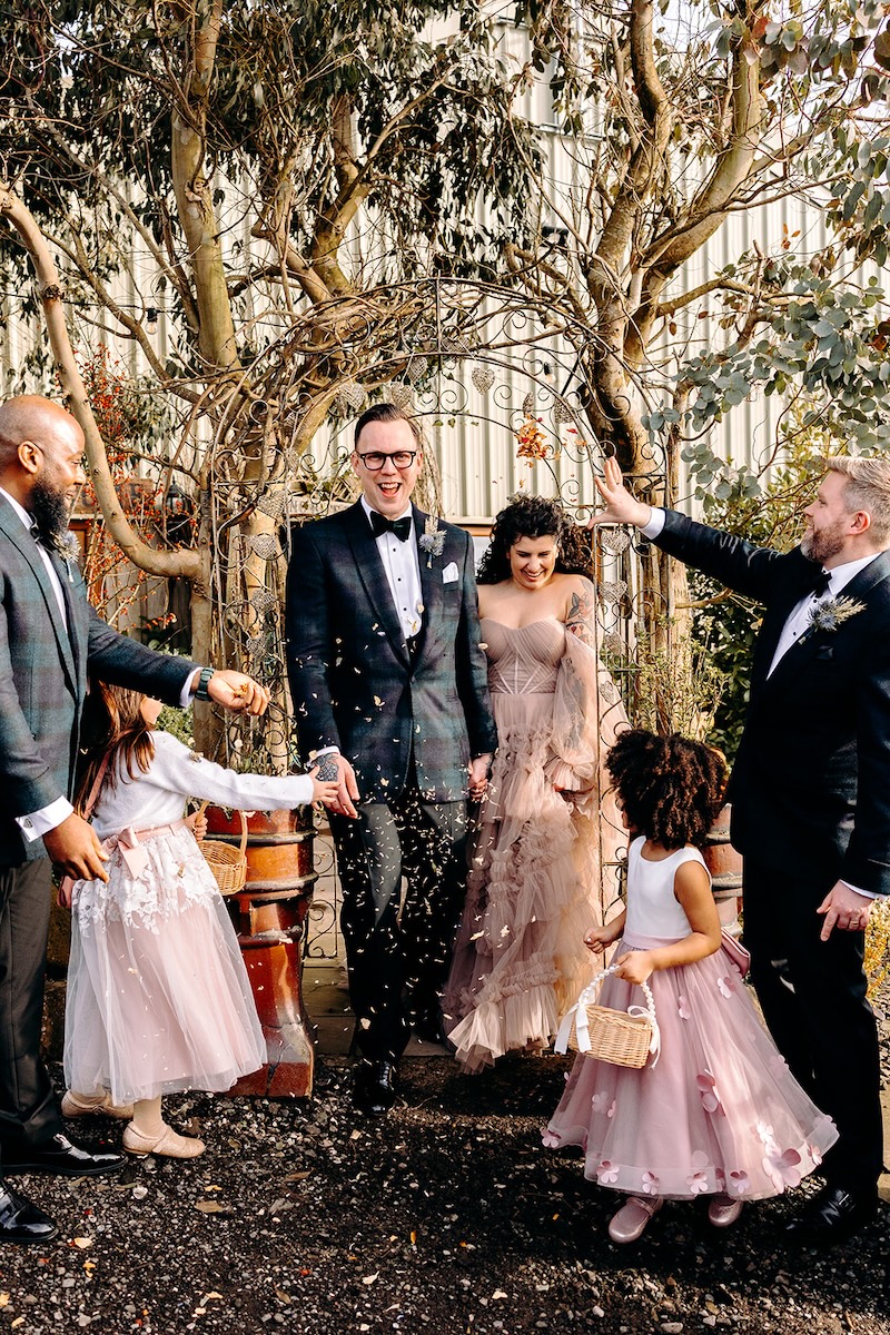 Bride and groom walking through confetti thrown by guests outside The Wellbeing Farm barn.