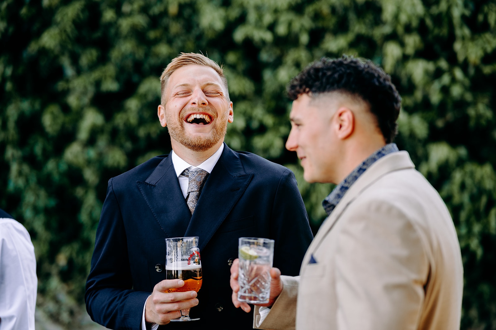 Two wedding guests laughing together over drinks during a relaxed Derbyshire wedding.
