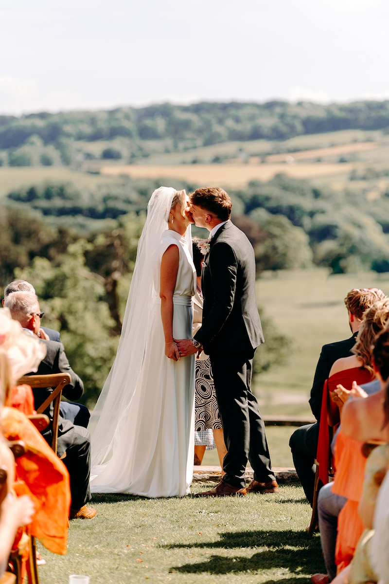 Newlyweds sharing their first kiss with Thornbridge Hall and wedding guests behind them.