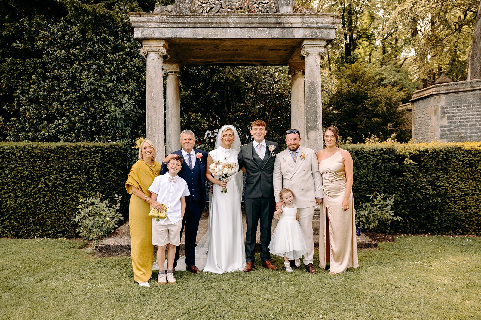 Family wedding photograph beneath a stone temple in the gardens at Thornbridge Hall.